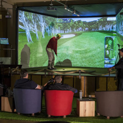 Bild vergrern: Blick auf den Indoorgolf-Simulator mit einem Spieler und Zuschauern
